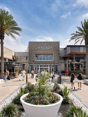 Centro comercial con restaurantes, fuente central, palmeras y personas caminando en un día soleado.